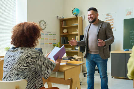 Smiling confident teacher in smart casualwear explaining subject to young female student with open...