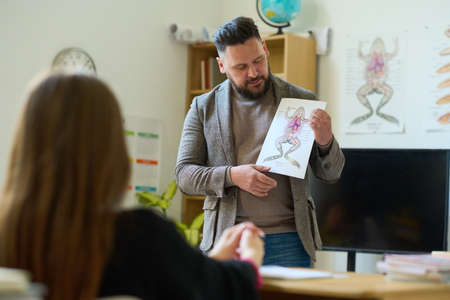 Confident mature biology teacher showing paper with frog picture to student while making...