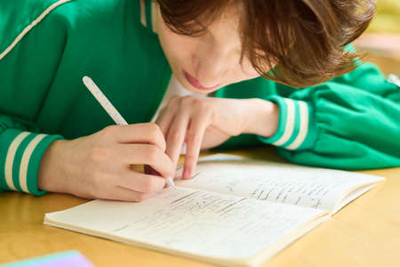 Teenage girl in green jacket bending over desk while making notes in copybook or writing down...