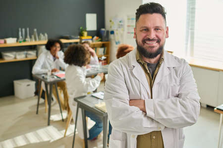 Happy bearded mature teacher of chemistry in labcoat crossing his arms on chest and looking at camera against group of teenage studentsの写真素材