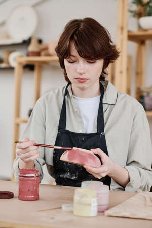 Young serious skilled female artisan with clay bowl and paintbrush painting handmade earthenware...
