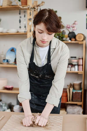 Young skilled female potter in apron bending over table while kneading clay on board before creating earthenware items for saleの写真素材