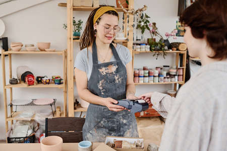 Young female owner of small earthenware shop with payment terminal selling handmade items to client paying for purchase by credit cardの写真素材