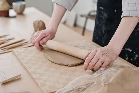 Hands of young craftswoman with wooden rolling pin flattening clay on board while working over new...