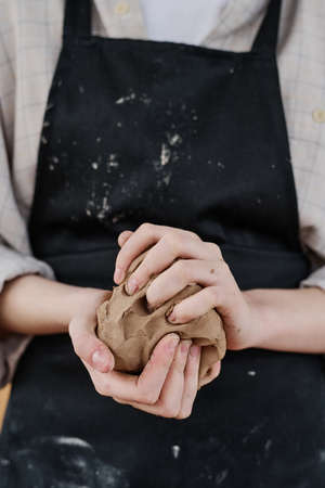 Hands of young female artisan in black apron kneading piece of clay for making earthenware or other creative clay items for saleの写真素材