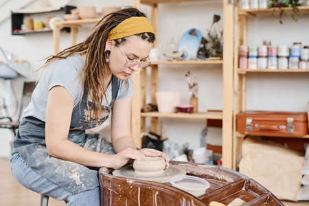 Young creative woman in casualwear and apron bending over pottery wheel and creating new earthenware item in workshopの写真素材