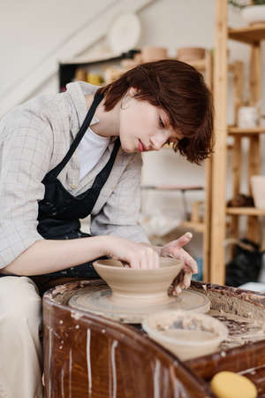 Young female pitcher bending over rotating pottery wheel during work over new clay item while...