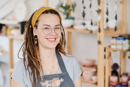 Happy young female clerk or owner of small shop looking at camera while standing against display with handmade earthenwareの写真素材