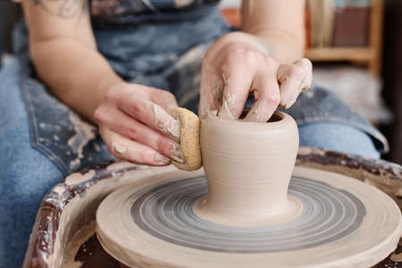 Hand of female potter holding sponge close to rotating clay item on pottery wheel while creating new earthenware in workshopの写真素材