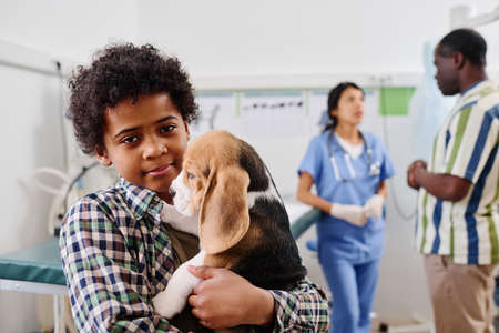 Selective focus of Black boy standing in vets office holding puppy, his grandpa and doctor talking...