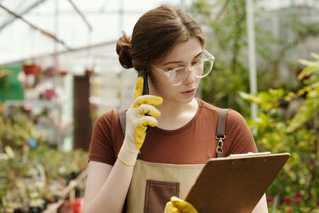Young female florist in eyeglasses checking the availability of flowers according to document while talking to customer on phoneの写真素材