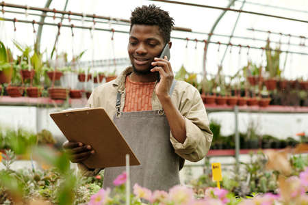 Flower shop owner looking at document in his hands and taking an order from customer by mobile phoneの写真素材