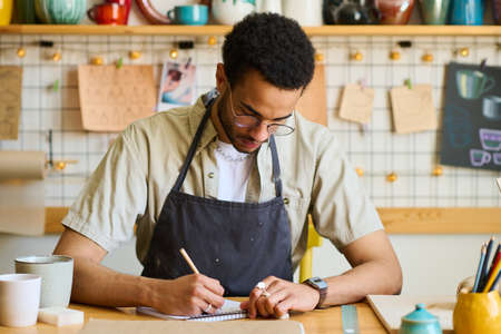 Young man in casual shirt and black apron drawing sketch of clay item in notepad while working over...
