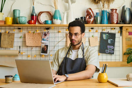 Young serious black man in workwear and eyeglasses sitting by workplace in front of laptop and...