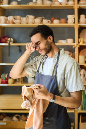 Young tired black man with towel touching his head after hard work over creating new handmade clay items for sale in workshopの写真素材