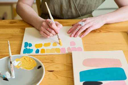 Hands of young woman with paintbrush making color swatches for her new painting while sitting by wooden table in workshopの写真素材
