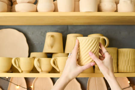Hands of young woman holding handmade clay mug while choosing one from assortment on shelves with other earthenware in shopの写真素材