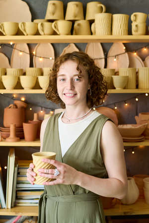 Young cheerful woman with handmade clay mug in hands standing by display with assortment of earthenware created for saleの写真素材