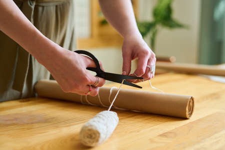 Hands of young female clerk cutting white cotton thread over rolled paper ordered by client before tying it to be prepared for sendingの写真素材