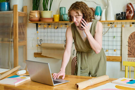 Young smiling female owner of handcraft shop bending over table with laptop and checking information...