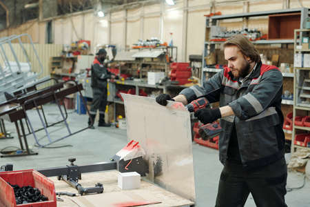 Young bearded male worker of warehouse or manufactory drilling metallic sheet while repairing part of industrial machineの写真素材