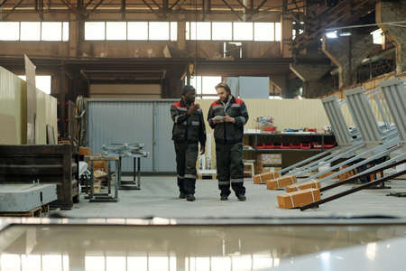 Two young intercultural male workers of factory having discussion of working points while moving along spacious workshopの写真素材