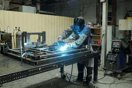 Young male worker of industrial plant bending over metallic workbench and welding parts of huge part...