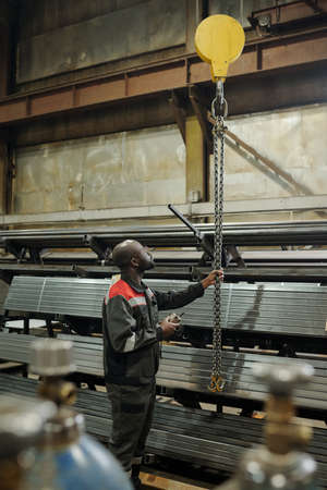Young black man pulling bunch of metallic chains while standing in front of stacks of materials for...