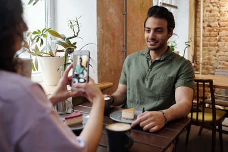 Young woman taking photo of her boyfriend with dark hair sitting by table in front of her and having...