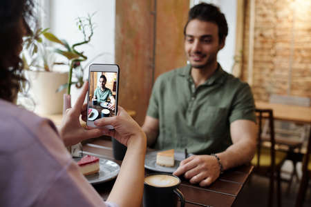 Hands of girl with mobile phone taking photo of her happy boyfriend sitting by table in front of her...