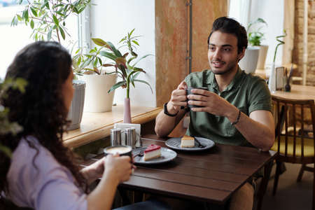 Guy with cup of coffee and his girlfriend sitting by table in front of one another and chatting while having coffee with tasty dessertの写真素材