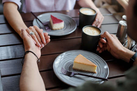 Hands of young affectionate dates sitting by table in front of one another while having coffee with yummy vanilla and blueberry cheesecakesの写真素材