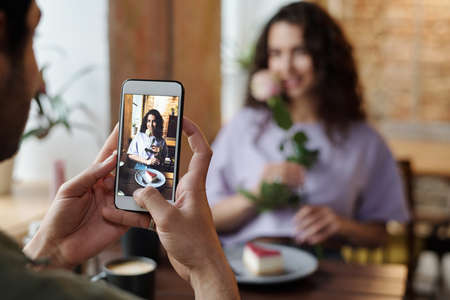 Hand of guy with smartphone taking photo of his girlfriend with rose and cheesecake sitting by table in front of him during romantic dateの写真素材