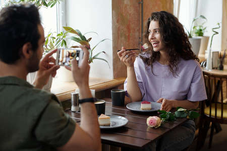 Happy young woman with piece of yummy cheesecake on spoon looking through window while posing for her boyfriend with smartphoneの写真素材