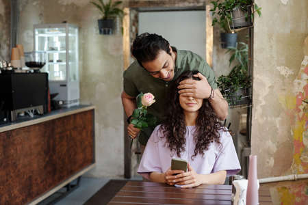 Young romantic man with pink rose covering eyes of his beautiful girlfriend with smartphone in hand...
