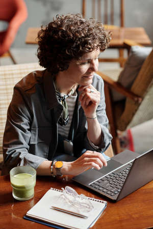 Happy middle aged businesswoman or freelancer networking by table in cafe while keeping hand on chin...