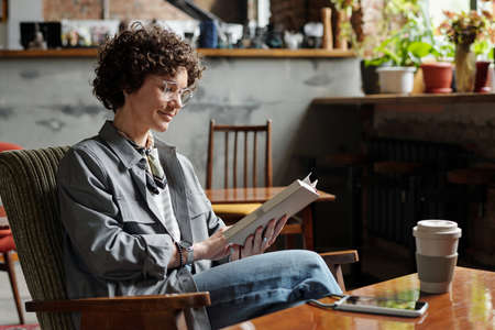 Happy young woman in casualwear and eyeglasses relaxing in armchair with interesting book while...