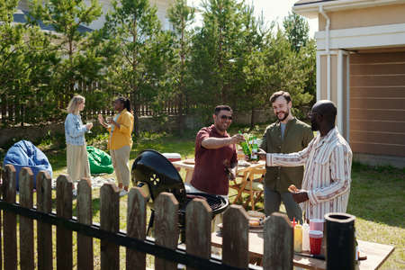 Three happy young intercultural men clinking with bottles of beer during outdoor party on weekend...