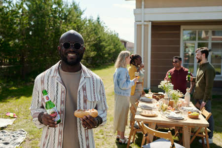 Young cheerful black man with bottle of beer and hotdog looking at camera during outdoor party...