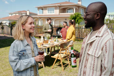 Happy young interracial friends or couple with drinks toasting and chatting during outdoor dinner against group of peopleの写真素材