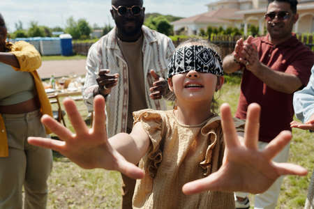 Cute blindfolded little girl playing hide and seek with young intercultural men and woman on...