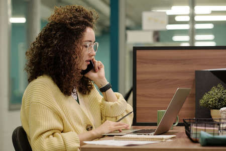 Side view of young serious businesswoman speaking to client on mobile phone and looking through online data on laptop screen in officeの写真素材