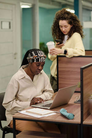 Two young intercultural businesswomen using mobile gadgets by workplace while looking for...