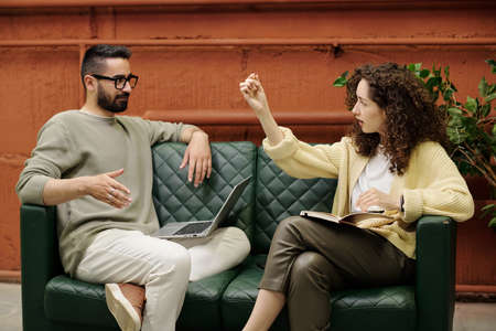 Confident female economist with notebook explaining analysis of financial data or development graph to colleague while both sitting on sofaの写真素材
