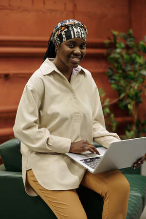 Young successful African American businesswoman with laptop on her knee networking and looking at...