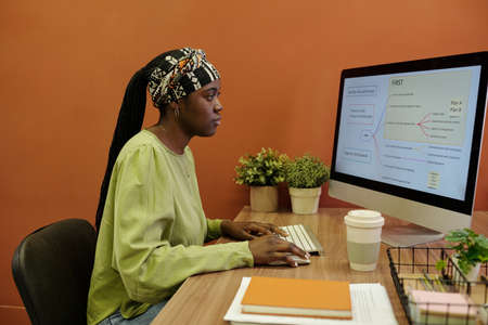 Side view of young black woman sitting by workplace in front of computer monitor and making scheme...