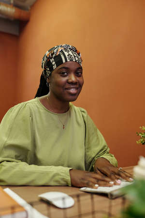 Happy young black woman in casual pullover and headband looking at camera by workplace in office...