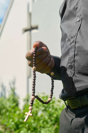 Hand of young African American priest with wooden rosary beads saying prayer or practicing silent pray in the garden by church buildingの写真素材