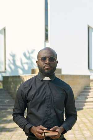 Young serious priest in black shirt with clerical collar holding Holy Bible while praying or pronouncing sermon in front of church buildingの写真素材