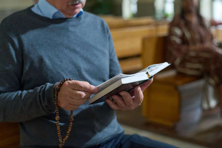 Hands of aged man holding open Bible and wooden rosary beads while reading verses from Gospel or some other book during sermonの写真素材